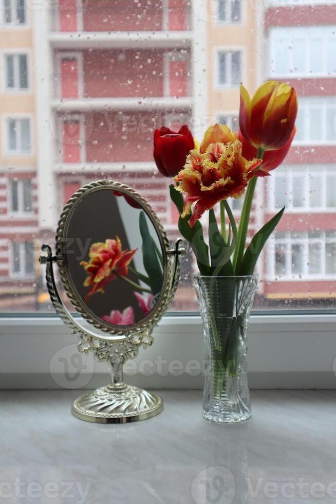 Bouquet of tulips and mirror with reflection of flower on the windowsill at rainy weather. photo