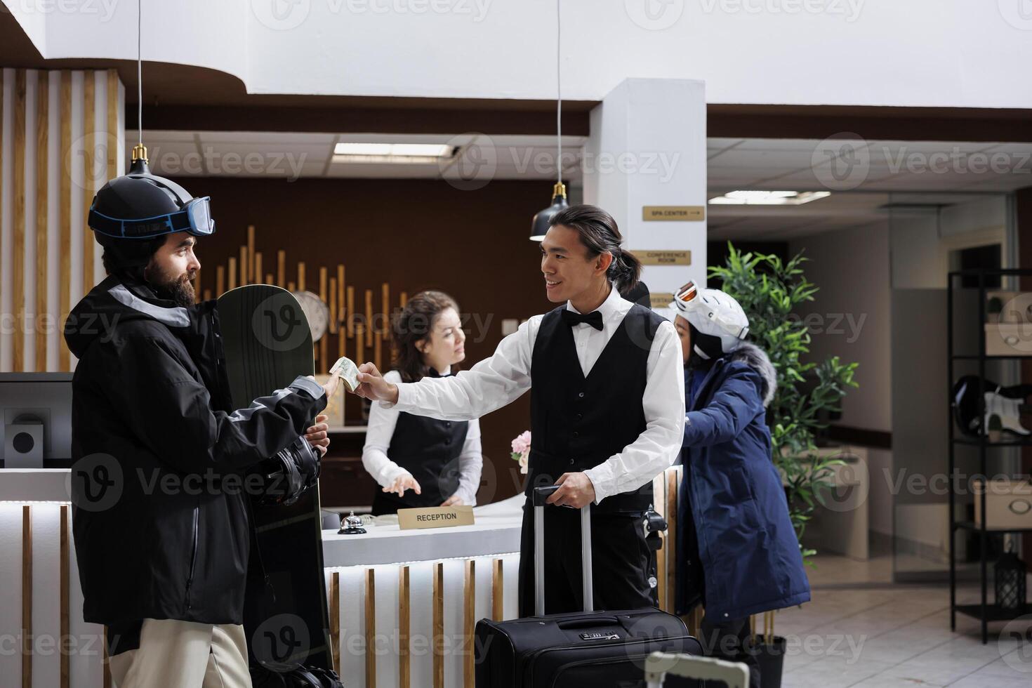 Tourists check in at ski mountain resort with luggage and snowboard equipment. Receptionist assists with reservations, male guest wearing winter clothing gives cash tip to staff in hotel lobby. photo