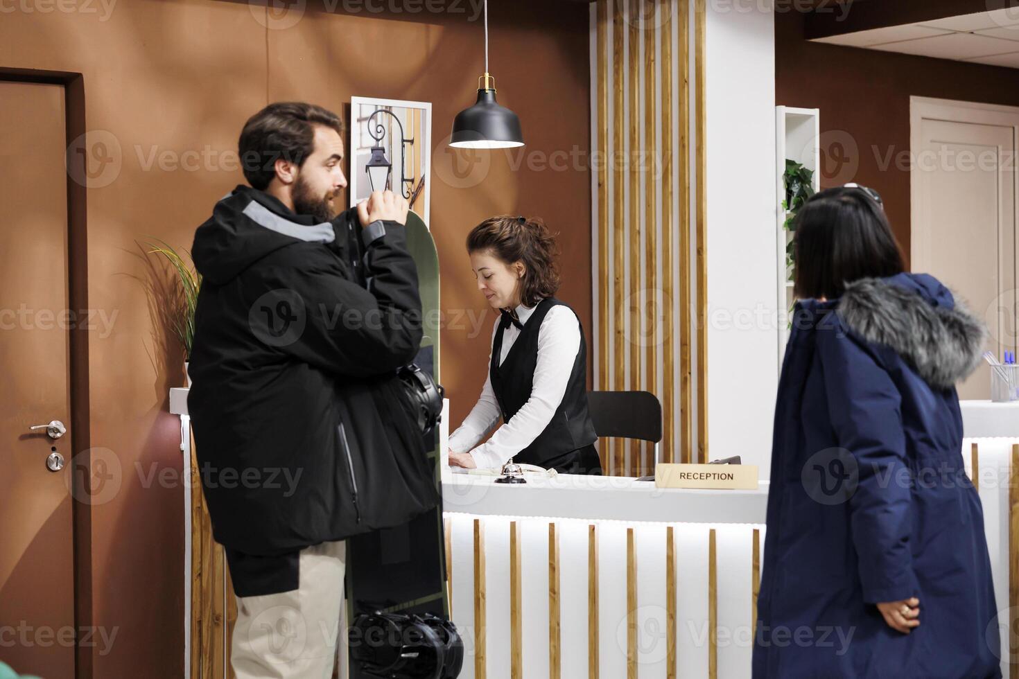Youthful man and woman at front desk being assisted with check-in by friendly hotel receptionist. Male tourist holding snowboard while female speaks with employee in resort lounge area. photo