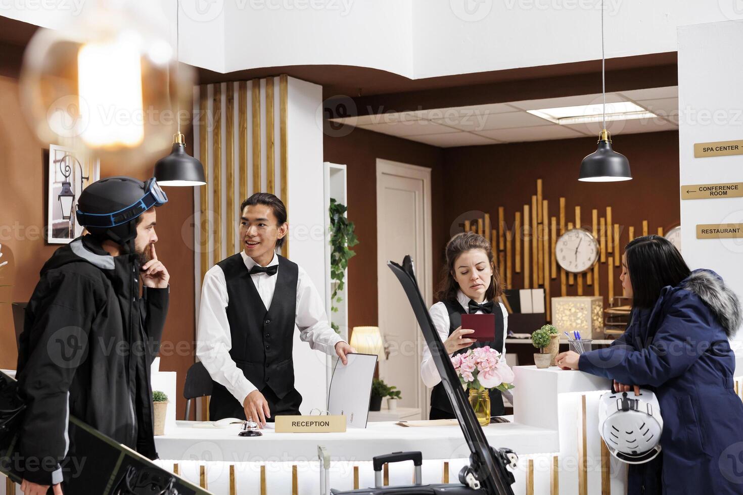 Visitors wearing snow clothing at counter of hotel reception doing registration process. Receptionists ensuring pleasant stay for guests with wintersports gear at exclusive ski mountain resort. photo