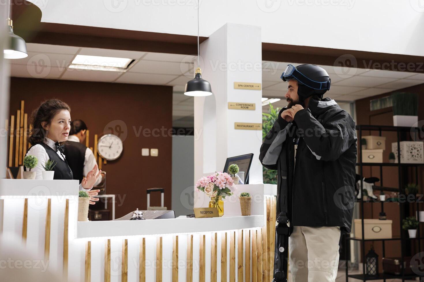 Caucasian man checking in at hotel reception for winter vacation at ski resort. Friendly female staff welcomes customer at front desk in cozy lounge area. Suitcases and ski gear in tow. photo