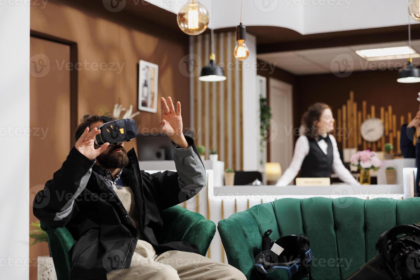 Male tourist wearing winter jacket is using a vr headset in lounge area of ski mountain resort. While waiting in hotel lobby man with beard utilizes vr goggles for research on wintersports activity. photo