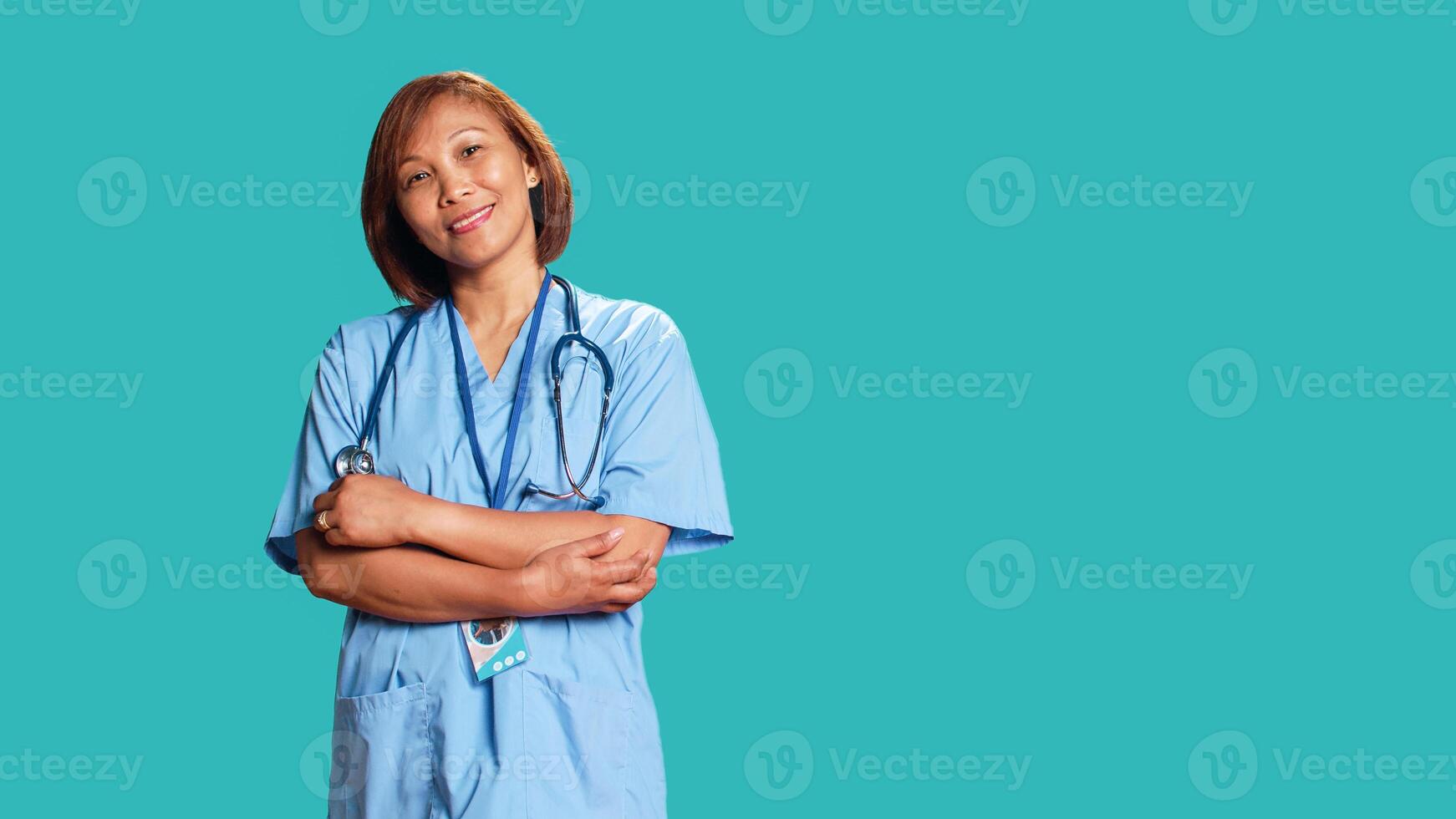 Joyful experienced asian nurse folding arms, smiling while looking at camera. Portrait of happy healthcare expert wearing protective scrubs, isolated over blue studio background photo