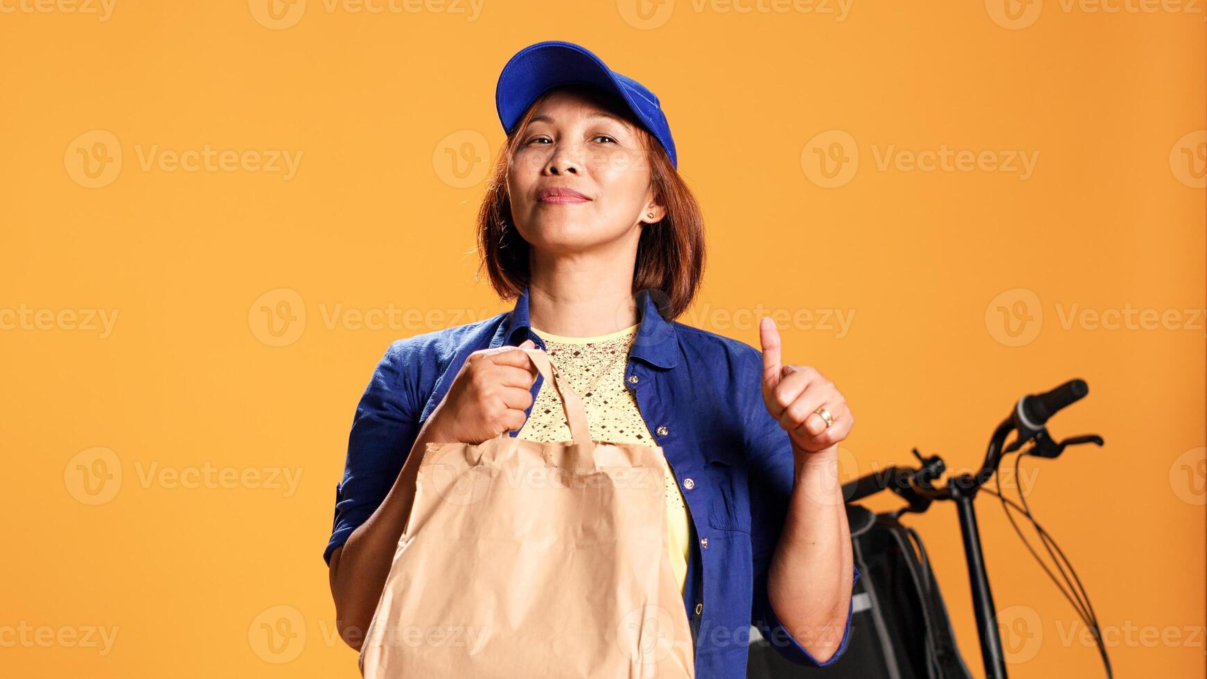 Slow motion close up portrait shot of courier waiting for customer to pick up food order. Bicycle rider delivering meal bag to client, showing approval gesturing, isolated over studio background photo