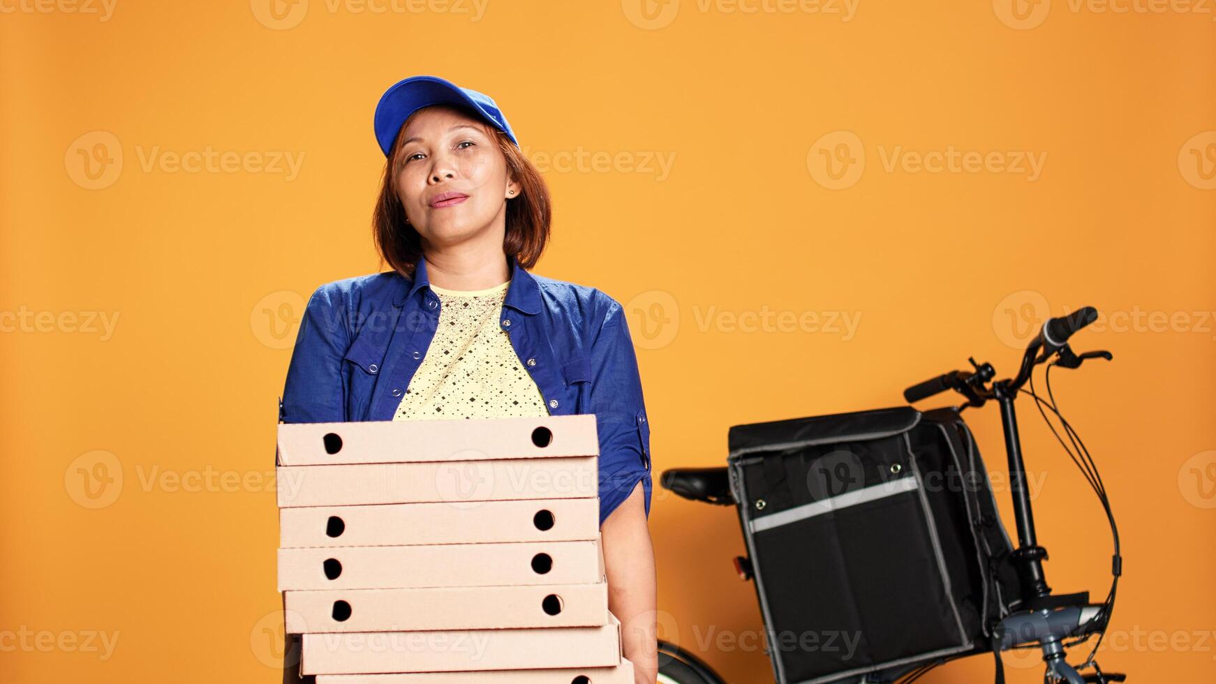 Impatient asian worker waiting for customer to answer door and pick up food. Portrait of bored courier isolated over orange studio background carrying pile of pizza boxes photo