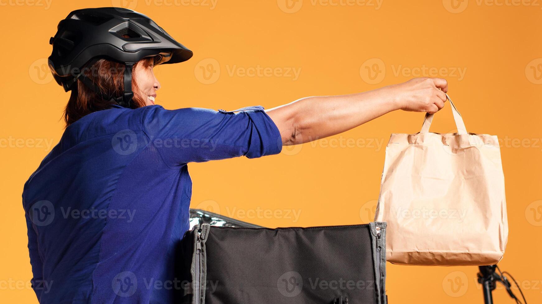 Close up shot of asian courier offering customer lunch bag order. Food delivery friendly worker arriving quick to client address, taking meal out of thermic backpack, isolated over studio background photo