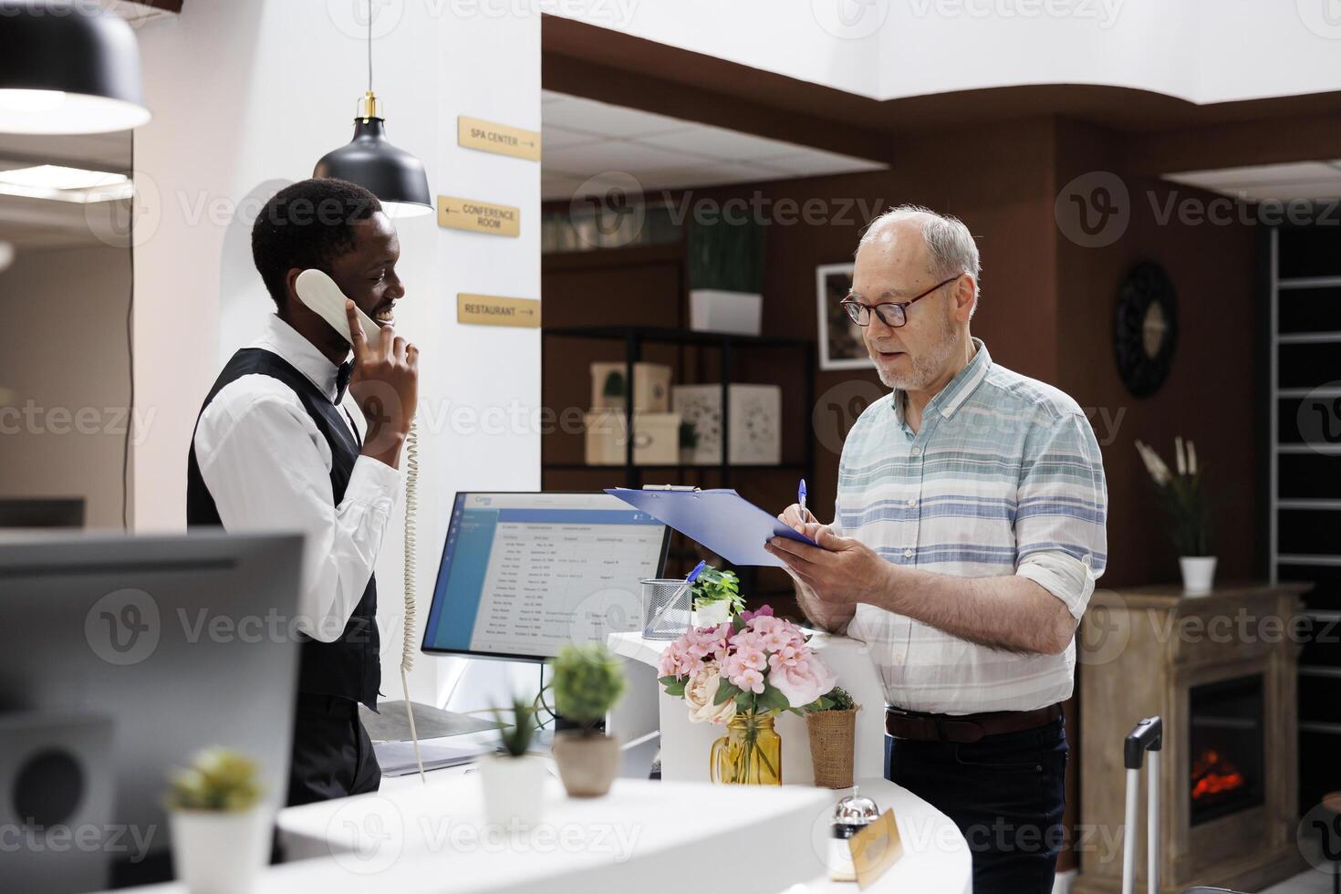 Caucasian retired old man signs reservation documents at hotel front desk as african american concierge answers call. Eldery male client filling check-in forms while receptionist speaks on telephone. photo