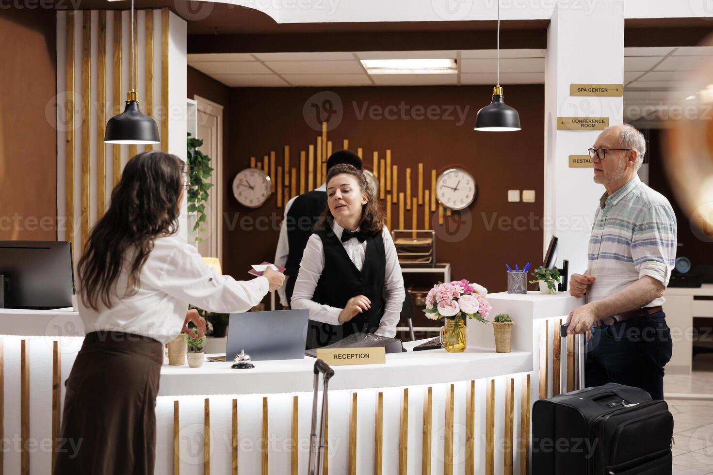 In hotel reception, elderly male and female travelers at registration counter awaiting reservation procedure. Retired senior woman giving personal documents to concierge for check-in. photo