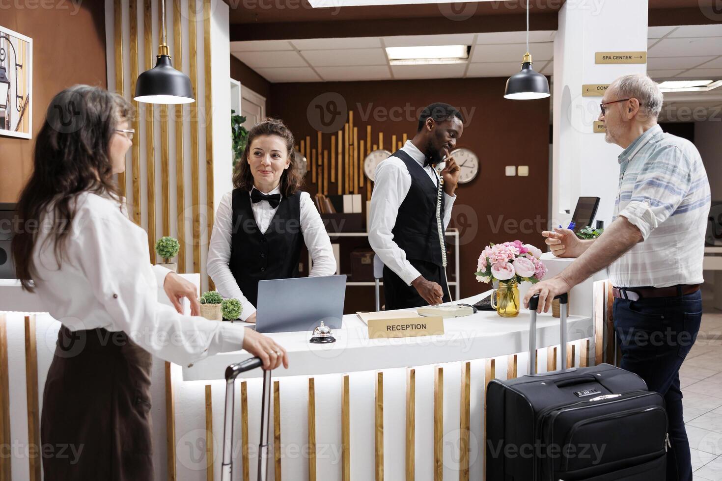 Two elderly travelers come at the hotel reception desk to check in. Multiethnic receptionists assist retired senior man and woman with booking and paperwork, therefore creating a pleasant environment. photo