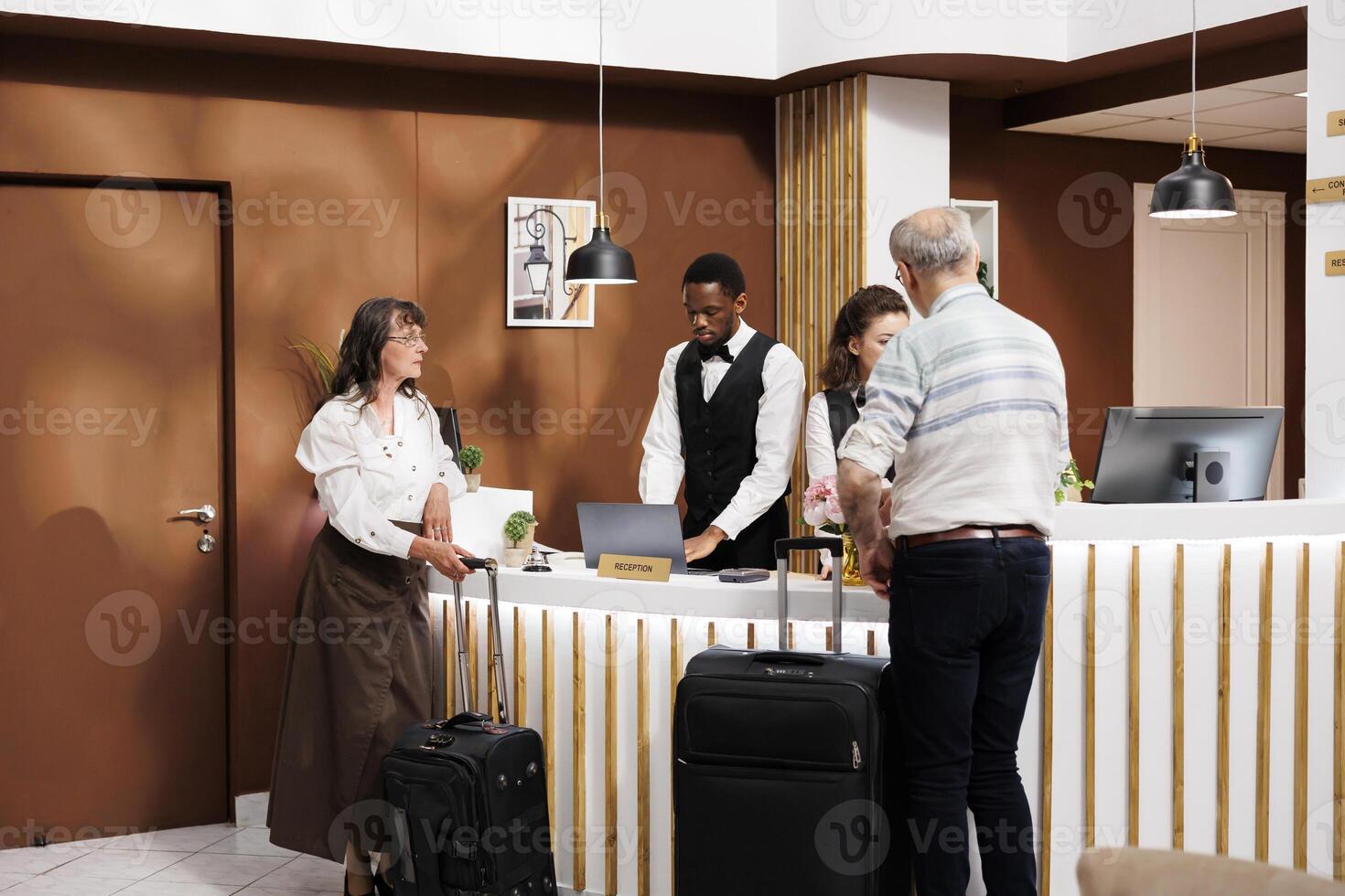 Male and female elderly guests in modern hotel lobby, with attentive staff providing efficient assistance during check-in and reservation procedures. Senior tourists wait with suitcases at front desk. photo