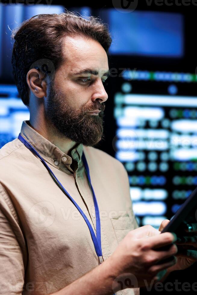 Programmer doing thorough system examination, checking data on tablet of needed debugging in server mainframes housing advanced data storage hardware supporting complex computer operations photo