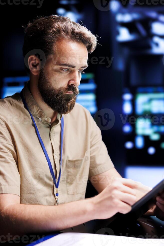 Repairman using tablet to examine server racks for power fluctuations, ensuring uninterrupted computing power. Worker monitoring data center, identifying potential vulnerabilities photo