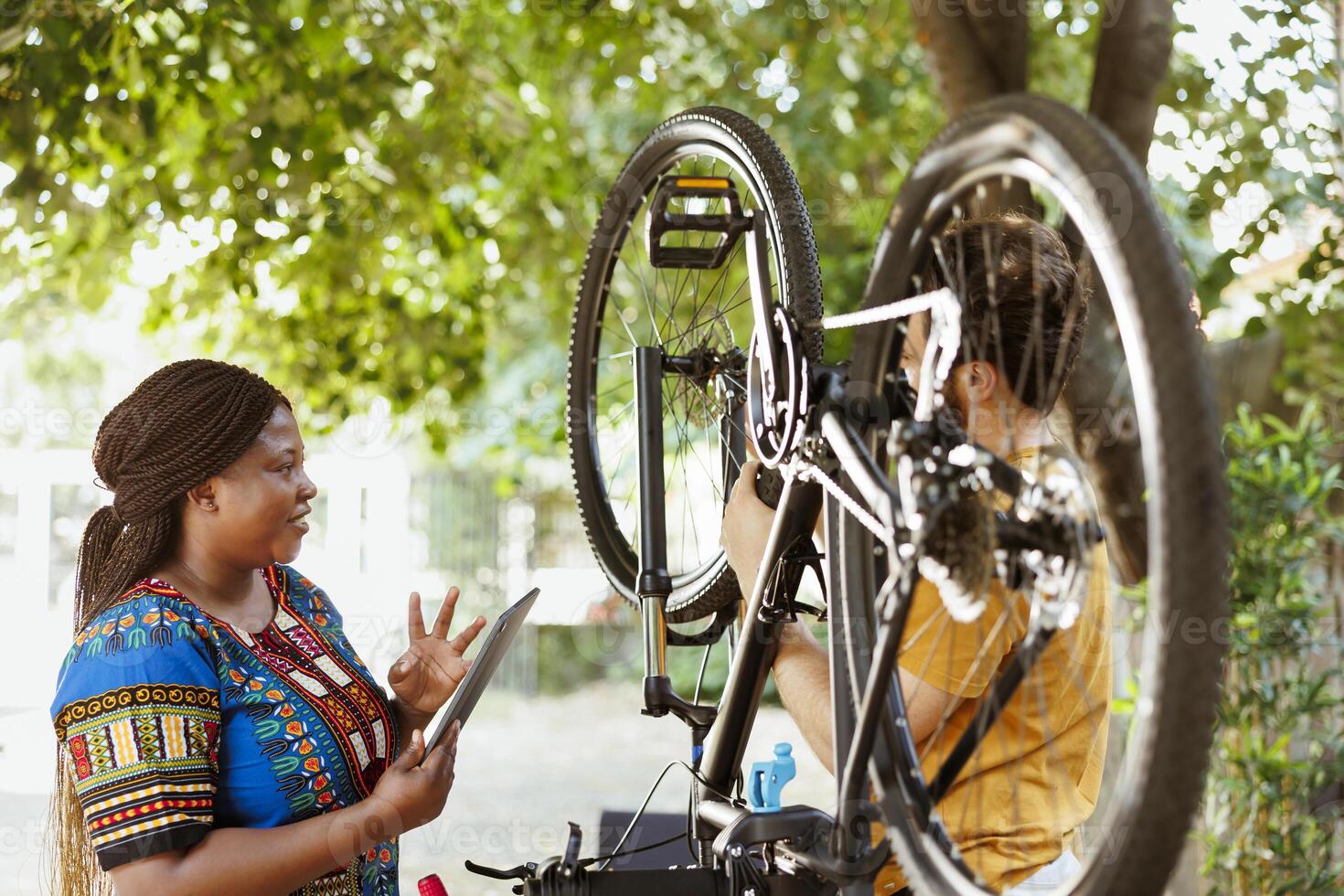 Vibrant black woman happily surfing internet to assist athletic man with bicycle maintenance. Smiling woman with digital tablet gives instructions to active young man to fix bike wheel and chain. photo