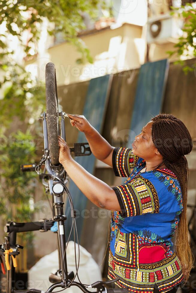 Using expert equipment, healthy young woman is repairing her own bicycle in yard as part of annual summer maintenance. Committed sporty african american lady fixes bicycle wheel with spanner as specialized work tool. photo