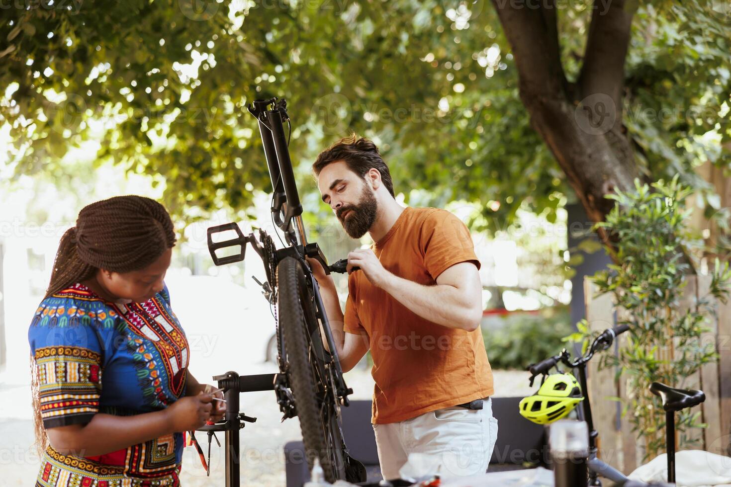Boyfriend and girlfriend repairing bicycle tire with work tools in yard for yearly maintenance. Multiethnic couple checking and fixing bike gear for summer cycling. photo