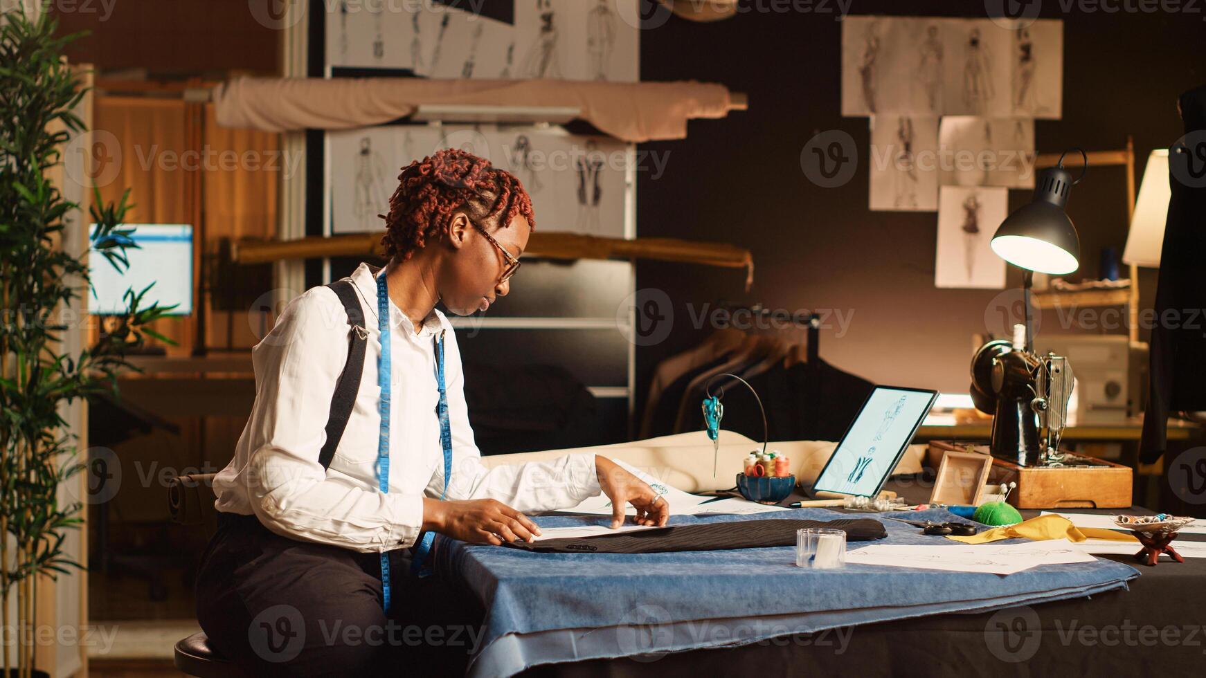 Female tailor designing clothes using sketches on tablet and papers, working on new fashion collection with modern design. Young couturier designing clothing line in tailoring atelier. Tripod shot. photo