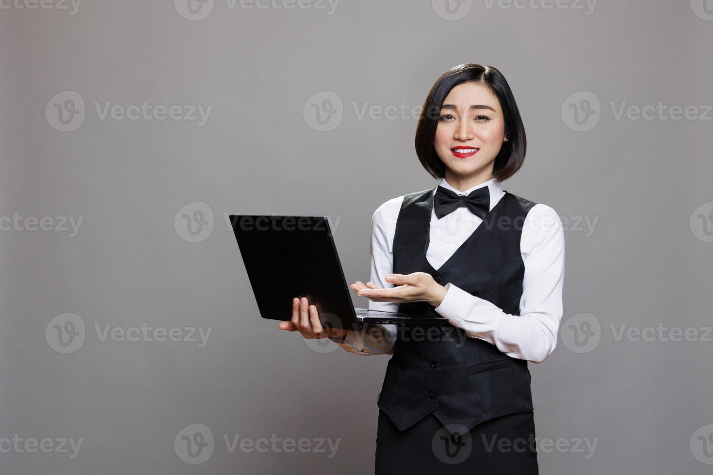 Smiling asian waitress in professional uniform showing laptop and looking at camera. Young attractive receptionist pointing with hand while holding portable computer portrait photo