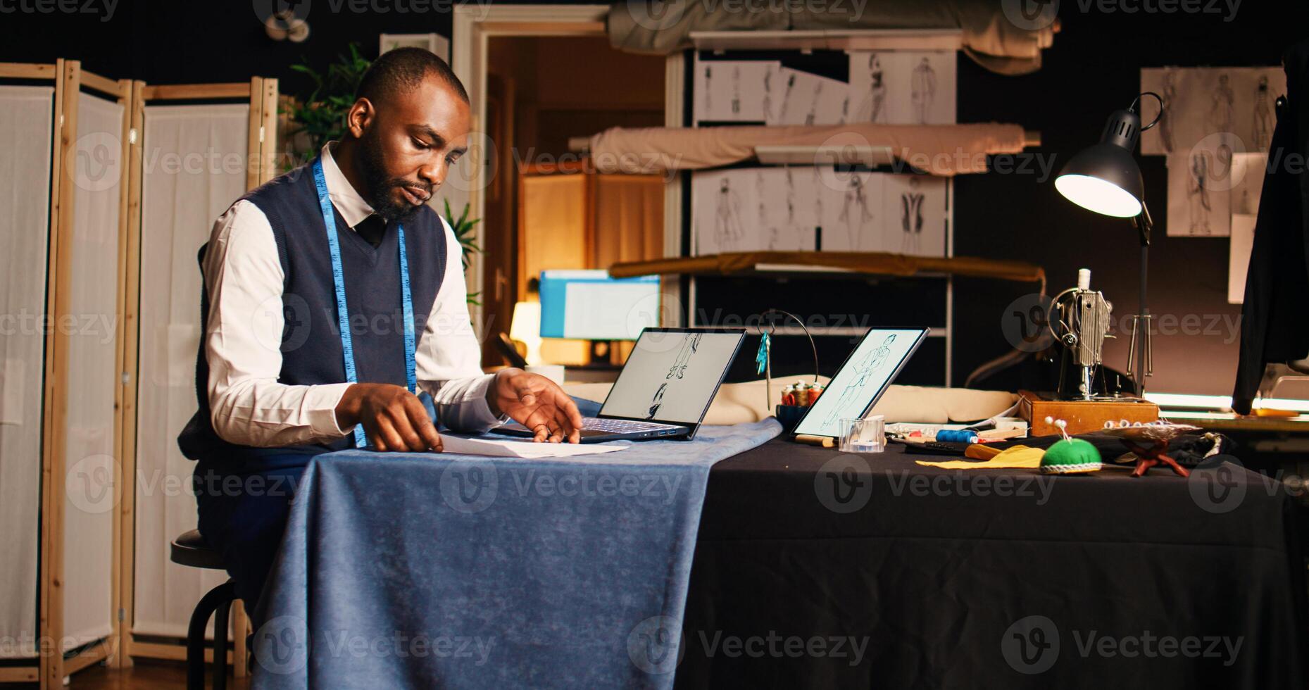 Professional couturier reviewing sketch, working with laptop and drawings to compare modern designs in atelier. Young tailor analyzing clothes models, needlework. Handheld shot. photo