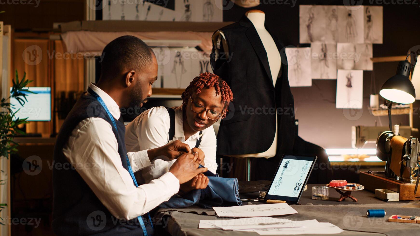 Two tailors choosing fabric for clothes in workshop, looking at materials on workstation before creating fashion collection. Team of couturiers crafting items with needlework. photo