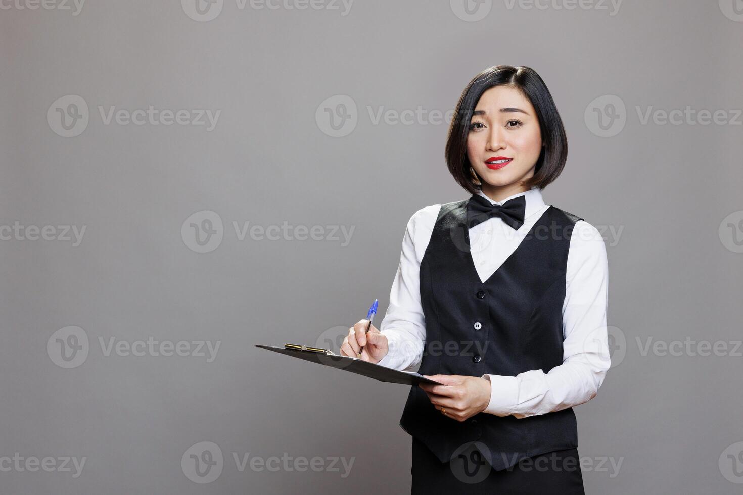 Young asian waitress in uniform holding clipboard, writing customer order and looking at camera. Attractive woman receptionist holding pen and checklist studio portrait on gray background photo