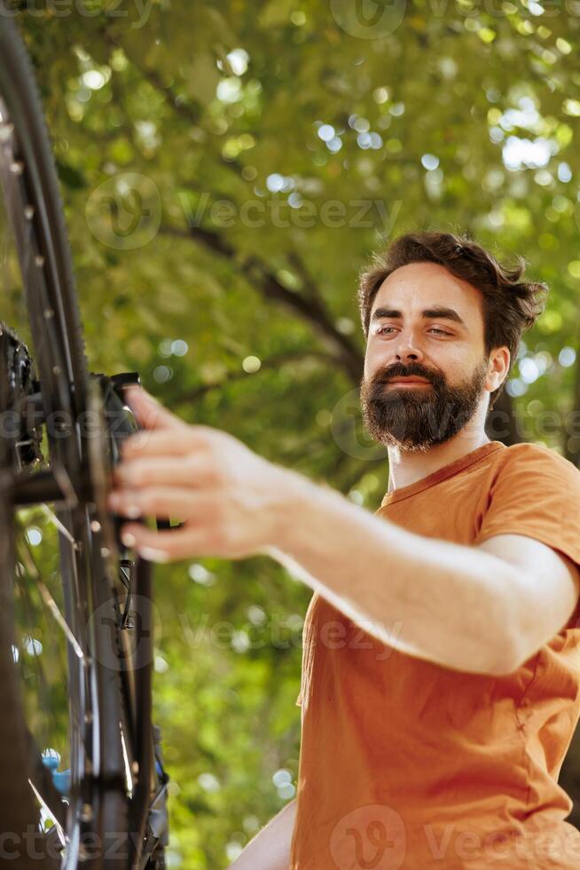 Close-up of young healthy caucasian man examining and adjusting bike wheel rubber and chain outside. Image showing enthusiastic sporty male working on bicycle chain ring in home yard. photo