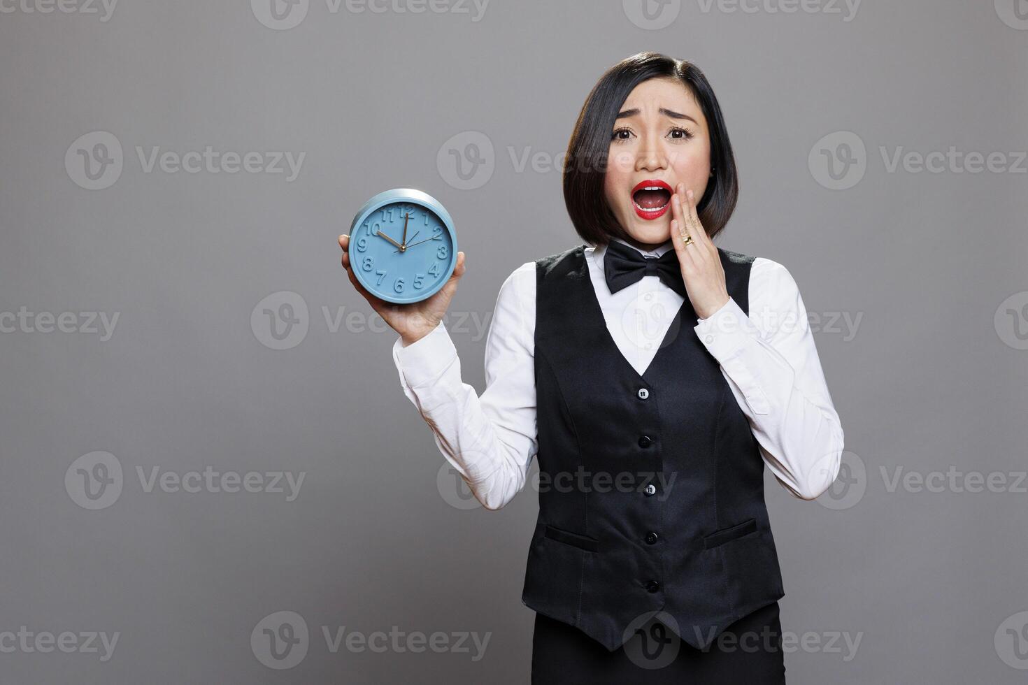 Anxious asian waitress panicking while holding circle dial alarm clock and looking at camera with open mouth. Young terrified woman receptionist showing timer watch portrait photo