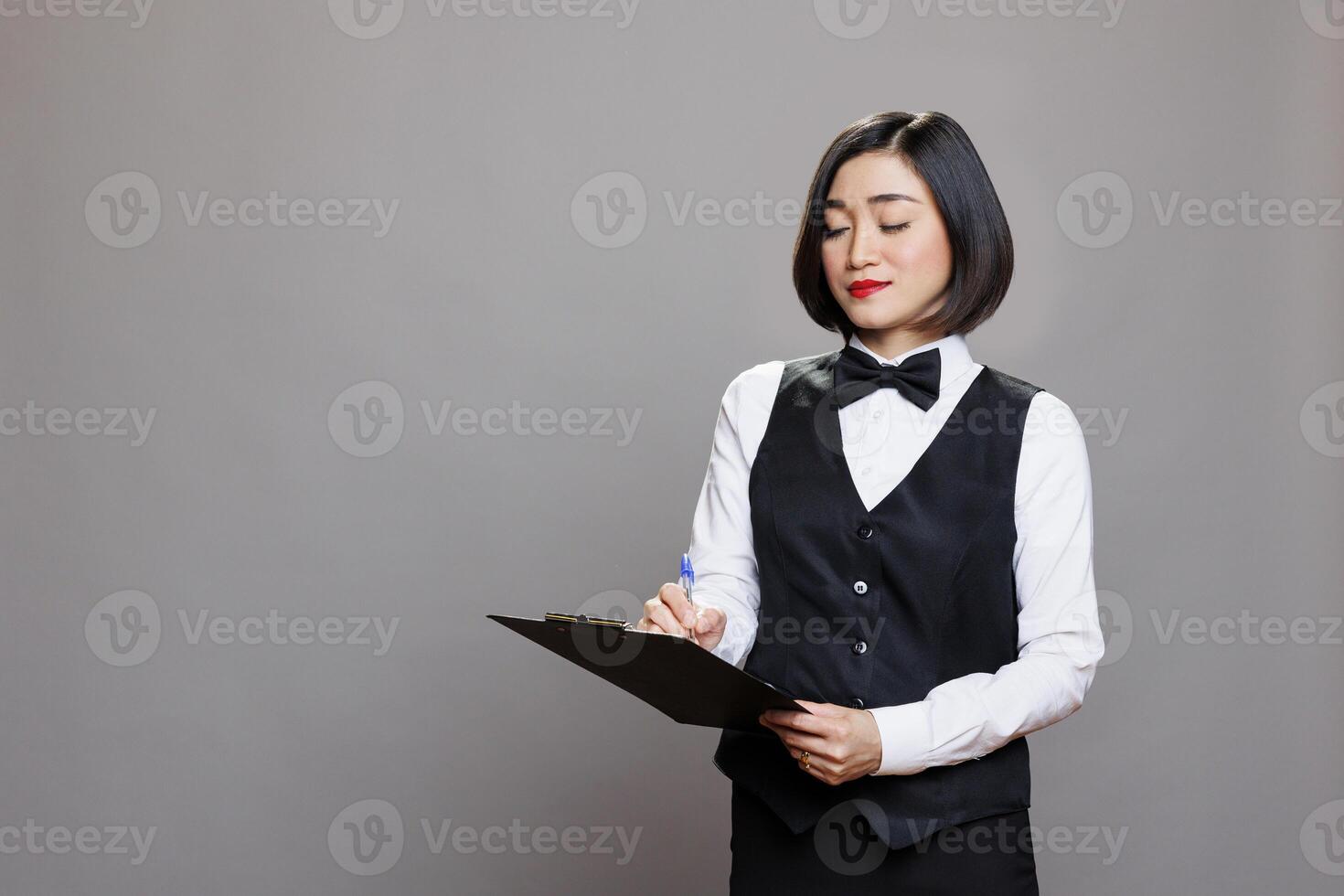 Asian waitress with focused expression writing on clipboard, checking list. Young attractive receptionist dressed in professional black and white uniform taking notes in studio photo