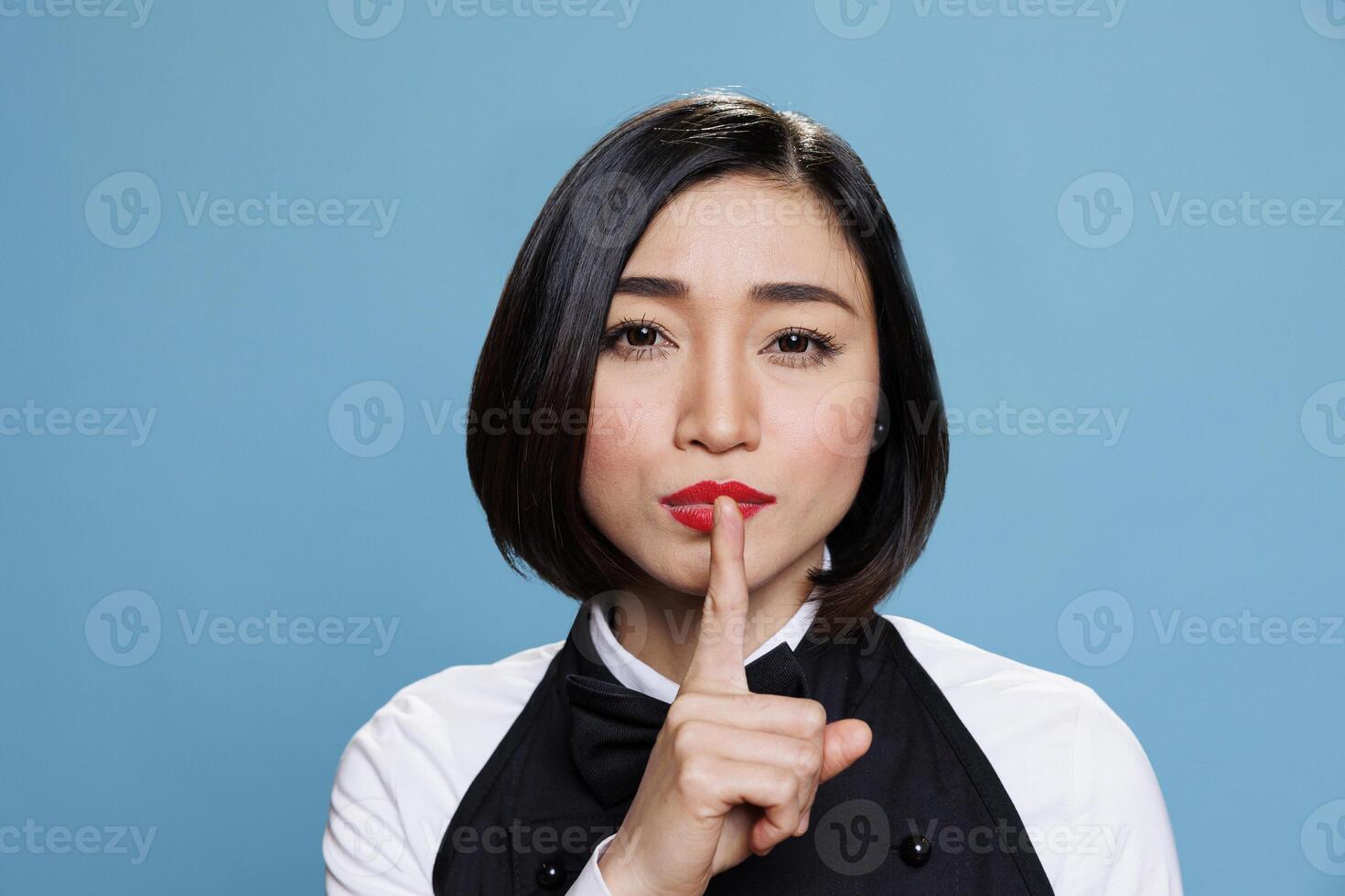 Hotel young asian woman receptionist making silent gesture, asking to keep secret portrait. Restaurant waitress wearing uniform holding forefinger on lips and looking at camera photo