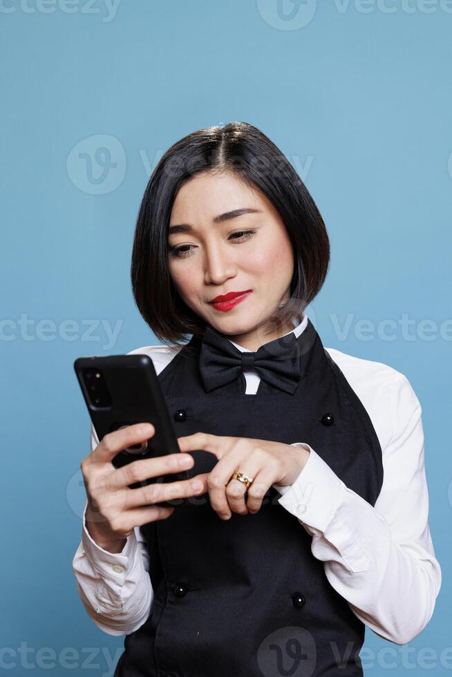 Young smiling asian waitress wearing restaurant uniform tapping on smartphone touchscreen. Attractive woman receptionist answering client message while using mobile phone photo