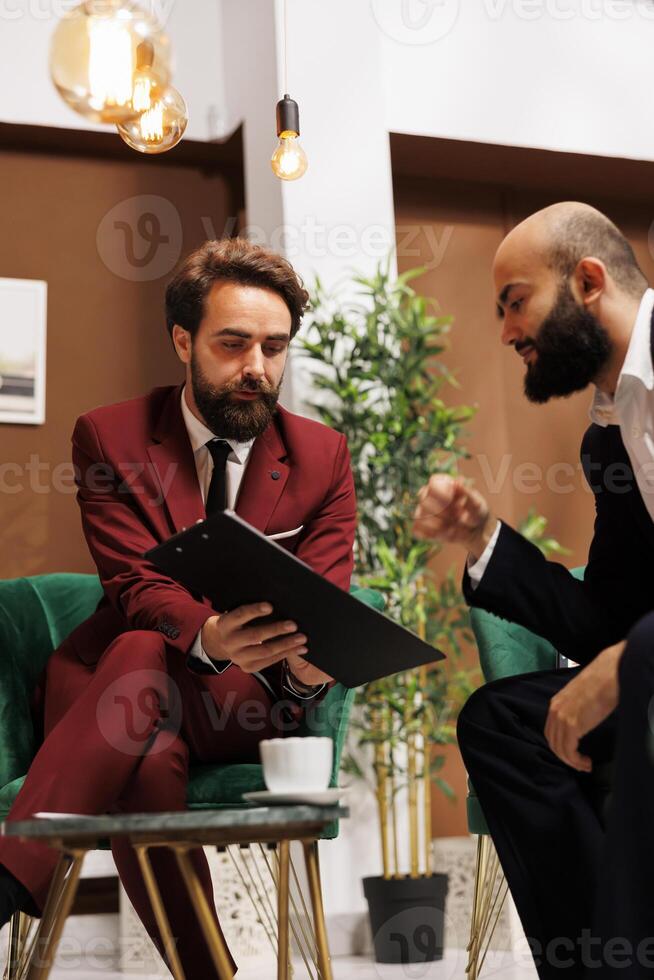 Management team review notes in lobby, preparing for upcoming international business meeting at luxury hotel. Two colleagues travelling for work trip, check conference speech on papers. photo