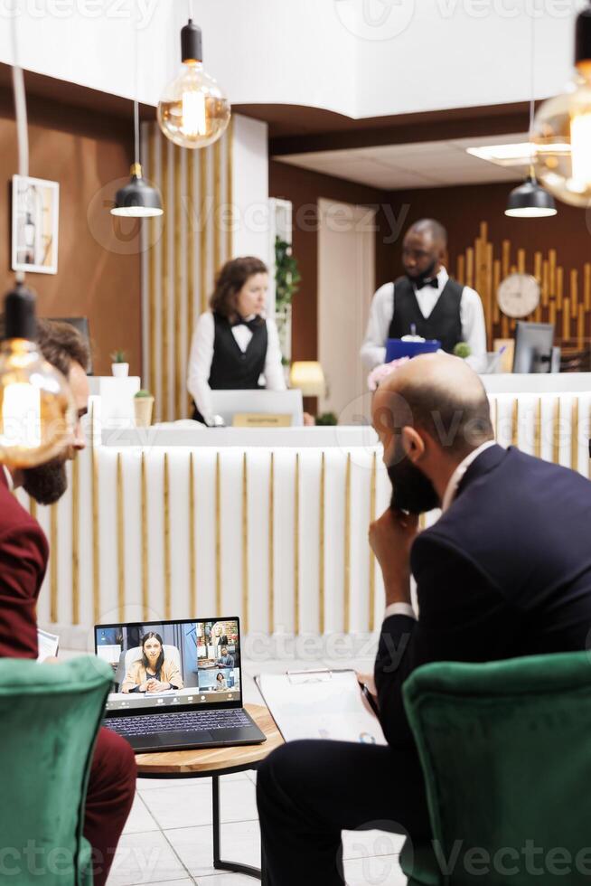 Entrepreneurs using laptop for online call, meeting clients in hotel reception lobby while they are travelling on work trip. Businessmen talking to people on remote videoconference chat. photo