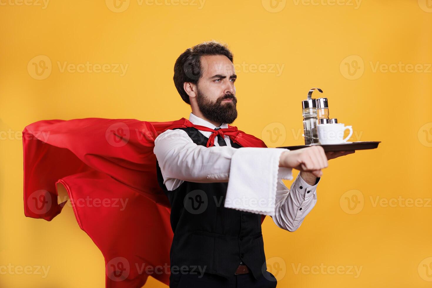 Elegant waiter with red cape carrying coffee on tray, having towel hanging over hand and posing as a superhero in studio. Young adult restaurant butler serving food and drinks. photo