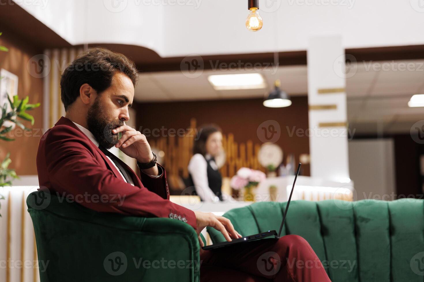 Company manager on business trip preparing to attend meeting in lounge area at hotel. Businessman waiting in reception lobby, checking presentation on laptop before starting conference. photo