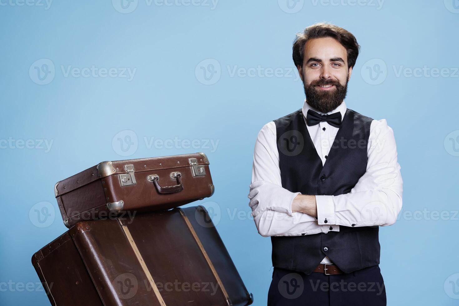 Hotel bellhop with luggage in studio, providing assistance to people and helping with trolley bags. Professional doorman or bellboy with suit and tie feeling confident on camera. photo