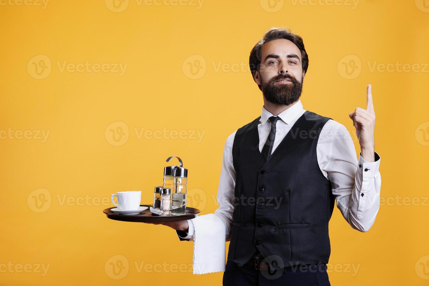 Waiter with serving tray points upwards on camera, showing something up above head while he wears professional restaurant clothes. Young man butler suggesting direction in studio. photo