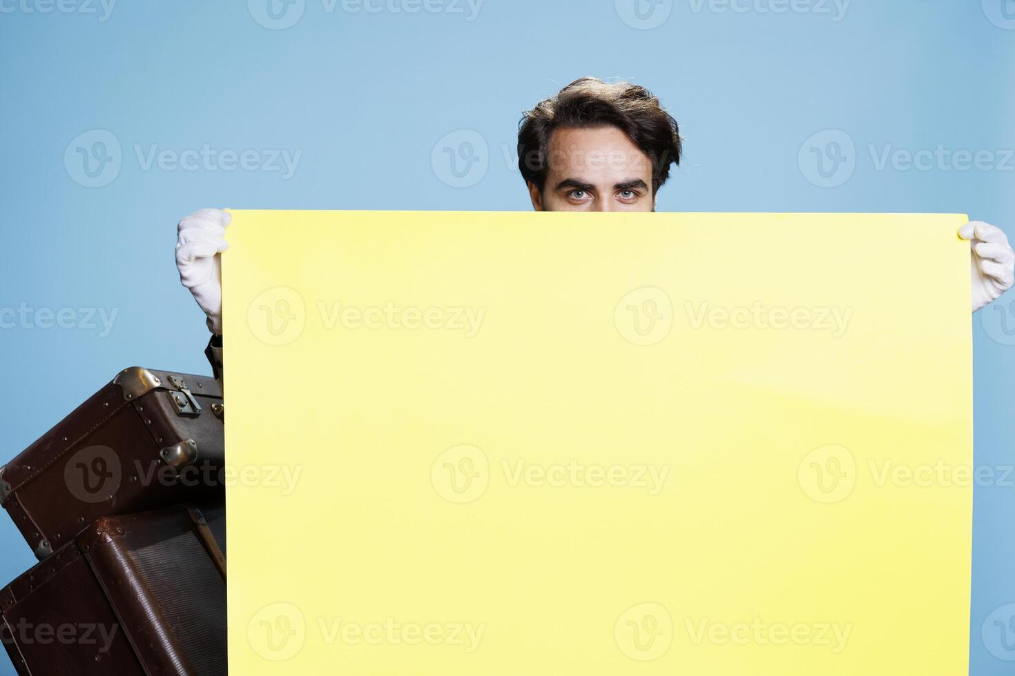 Doorkeeper presents empty ad banner in studio, young man works as hotel porter. Classy elegant bellboy showing blank yellow poster to do advertisement sign on camera, travel industry. photo