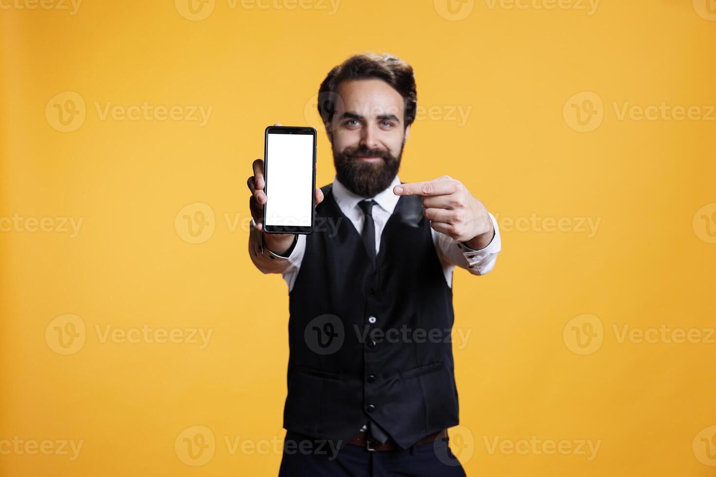 Confident waiter points to white screen on camera, presenting isolated mockup blank display on mobile phone. Young butler with suit and tie shows empty device with copyspace template. photo