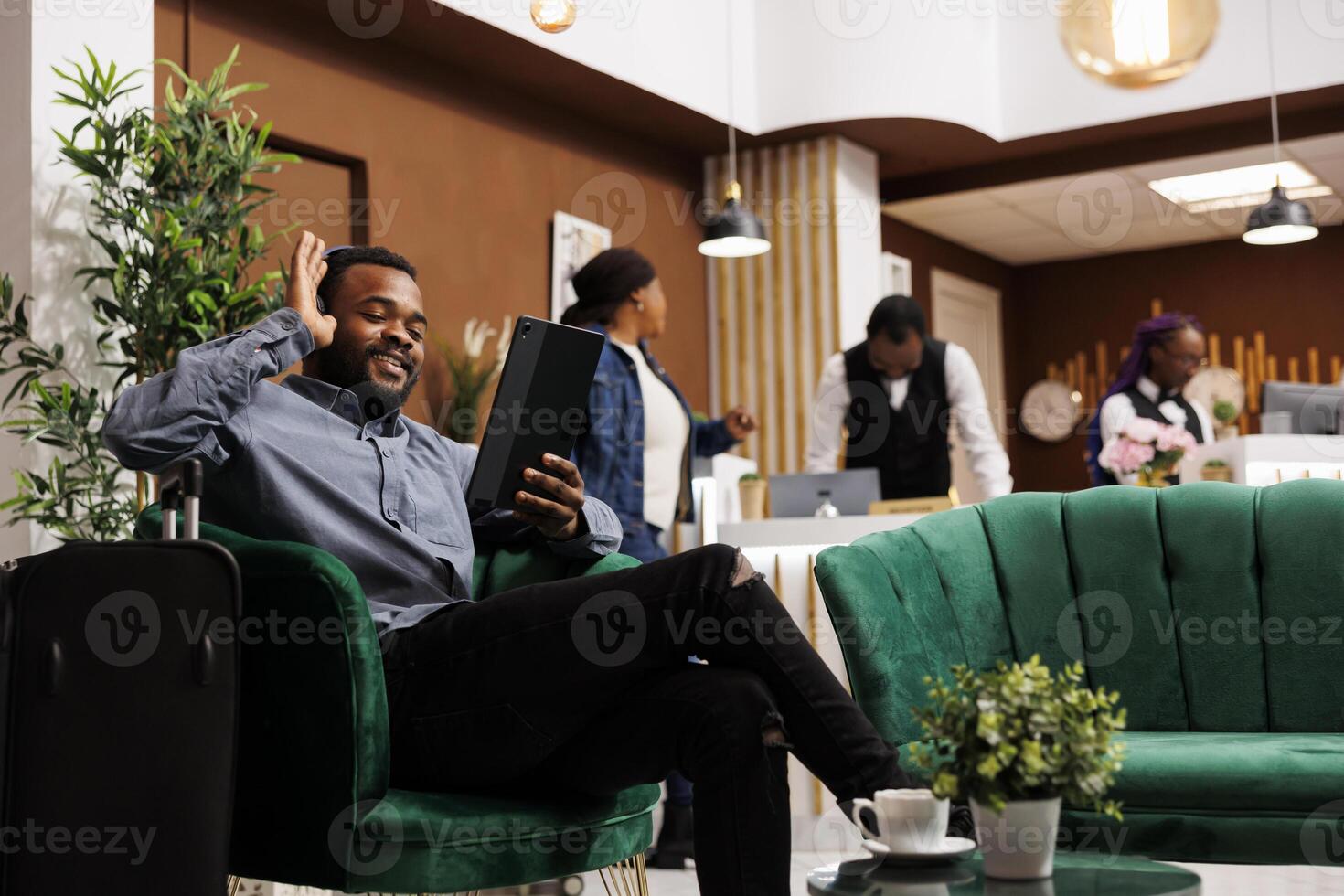Happy smiling young African American man traveler sitting in hotel lobby having call while waiting for check-in. Tourist holding digital tablet, waving hand greeting friend looking at camera photo