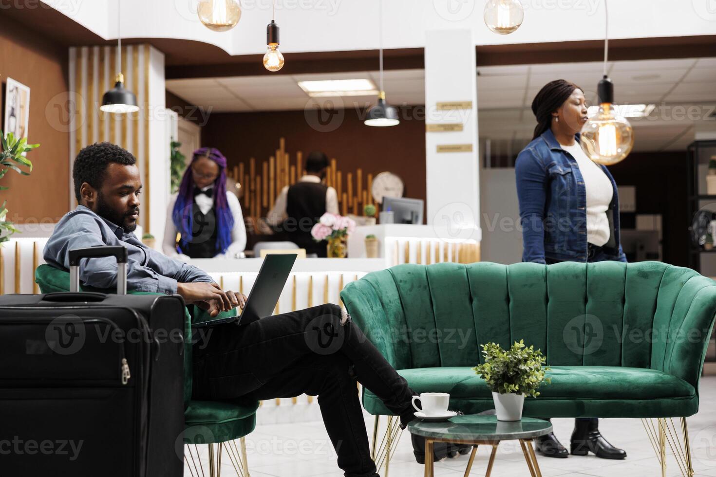 Busy African American man travelling freelancer sitting in armchair with suitcase and laptop at hotel lobby. Black millenial guy using computer working on project remotely during business trip photo