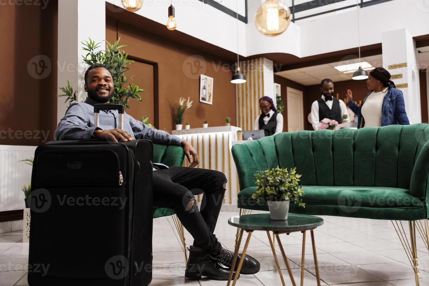Happy smiling African American man guest sitting in hotel lobby with suitcase awaiting for room. Positive millennial black guy tourist resting in armchair while waiting for check-in time photo