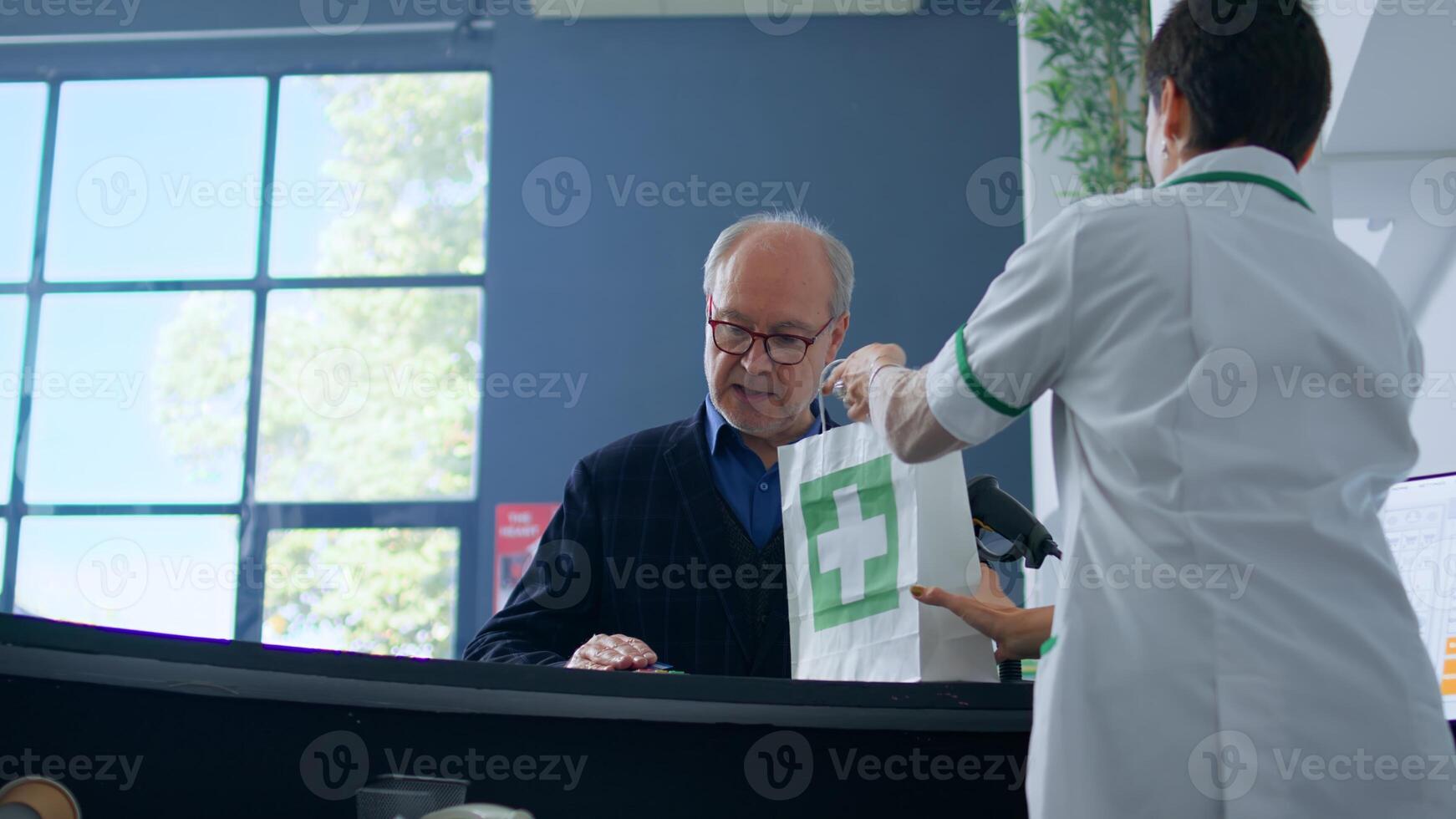 Elderly shopper arriving at drugstore checkout counter after finding needed medical items, purchasing pills with credit card, being helped by chemist assistant with shopping bag photo