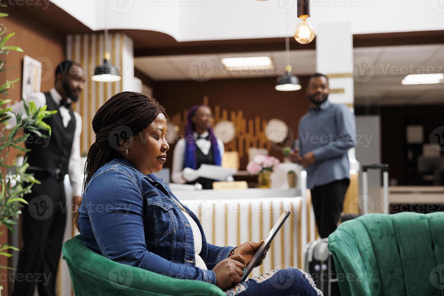 African American woman traveler sitting in luxury hotel lobby looking at tablet screen using digital device to check-in and receive room key. Female guest resting in lounge area of resort photo