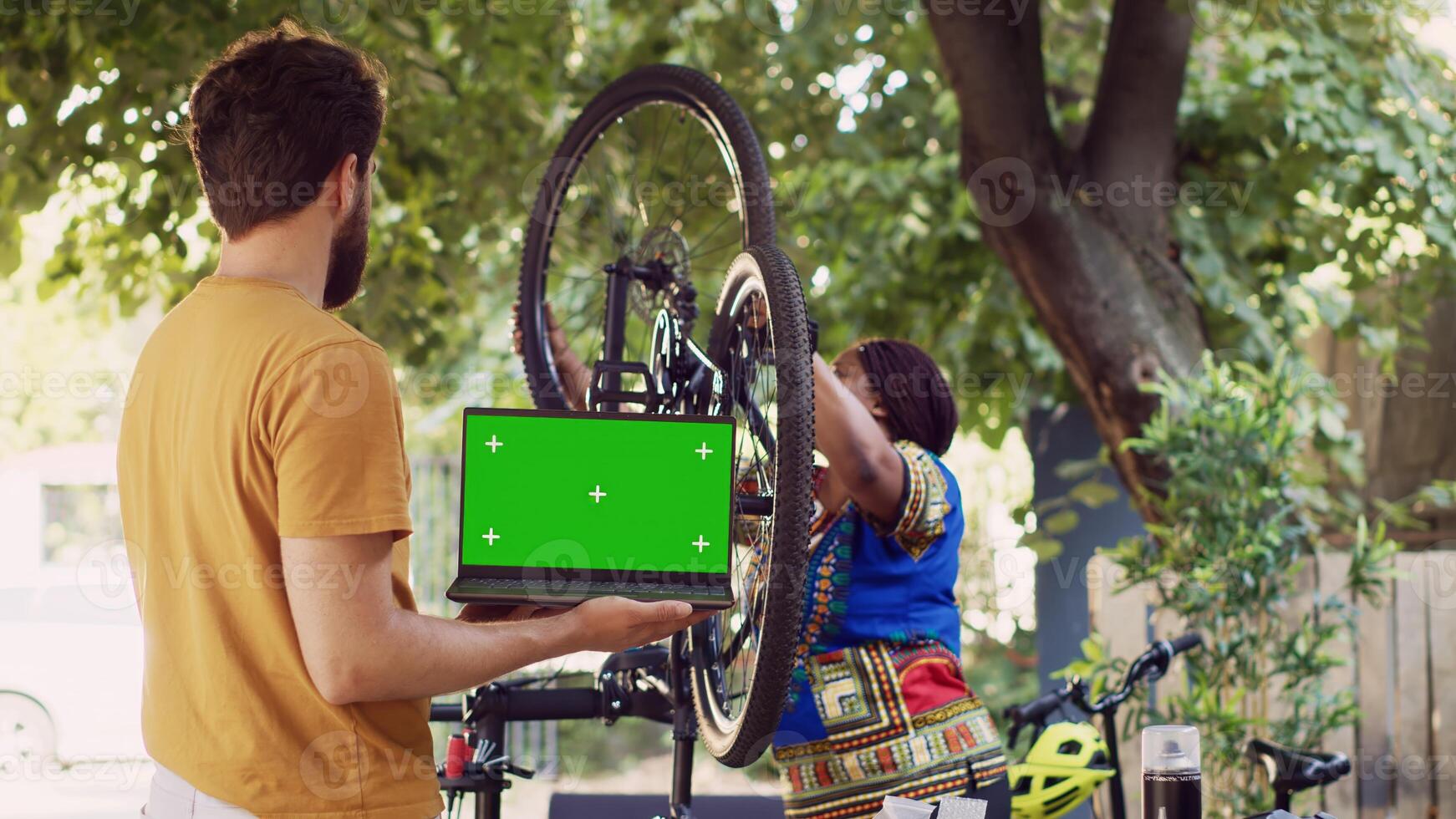 Young man with laptop displaying greenscreen as african american lady attaches wheel to bicycle frame in yard. Personal computer with isolated chromakey template held by caucasian male. photo