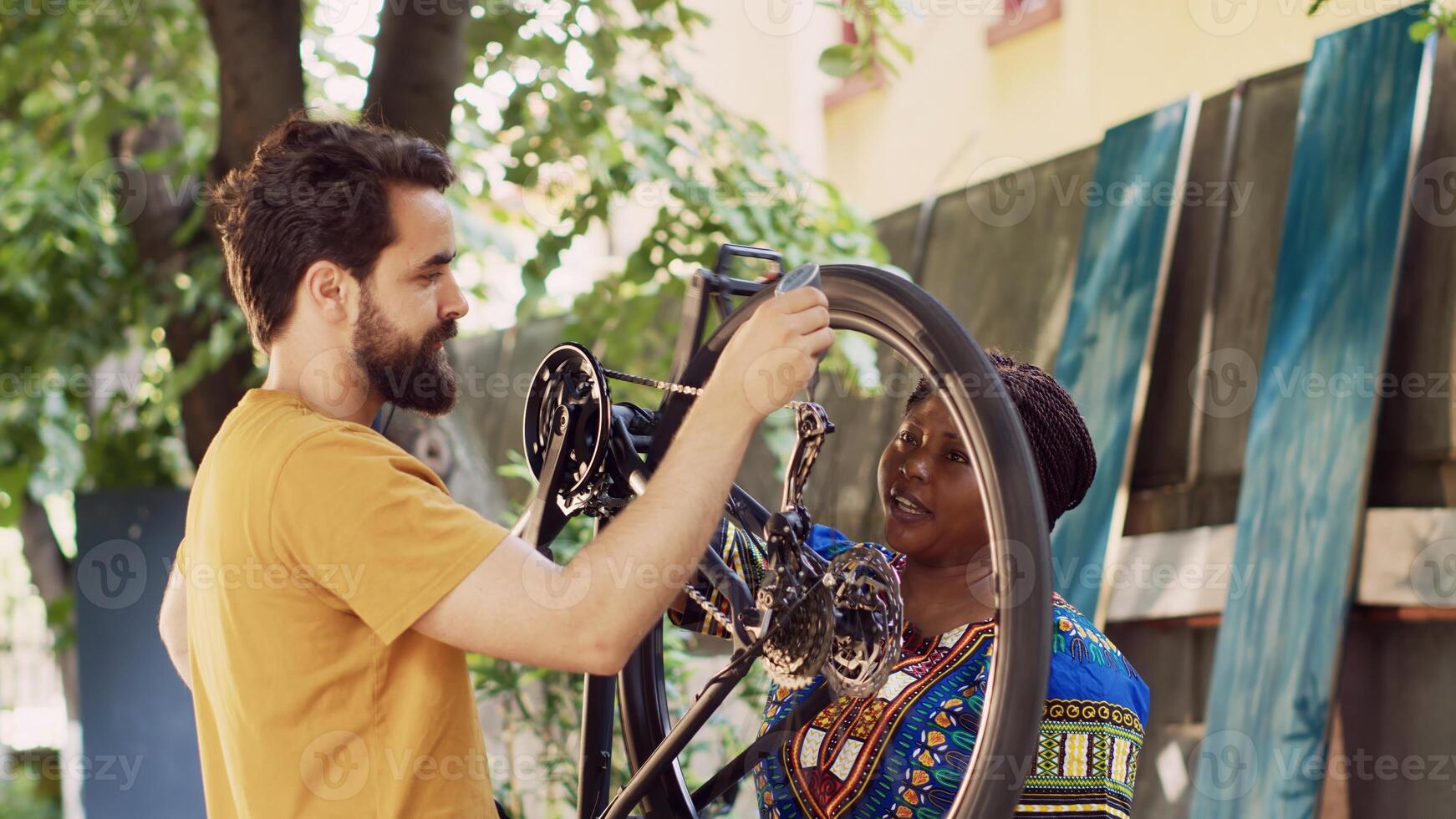Sporty multiethnic couple doing bike maintenance in home yard. Young african american woman grasping modern bicycle while healthy caucasian man diligently lubricating wheels outdoor. photo