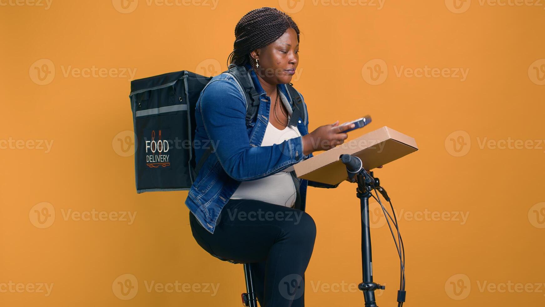 Black woman delivers takeout orders on bicycle, utilizing wireless payment device for seamless transactions. African american delivery person taking pos machine from pocket for contactless pay. photo