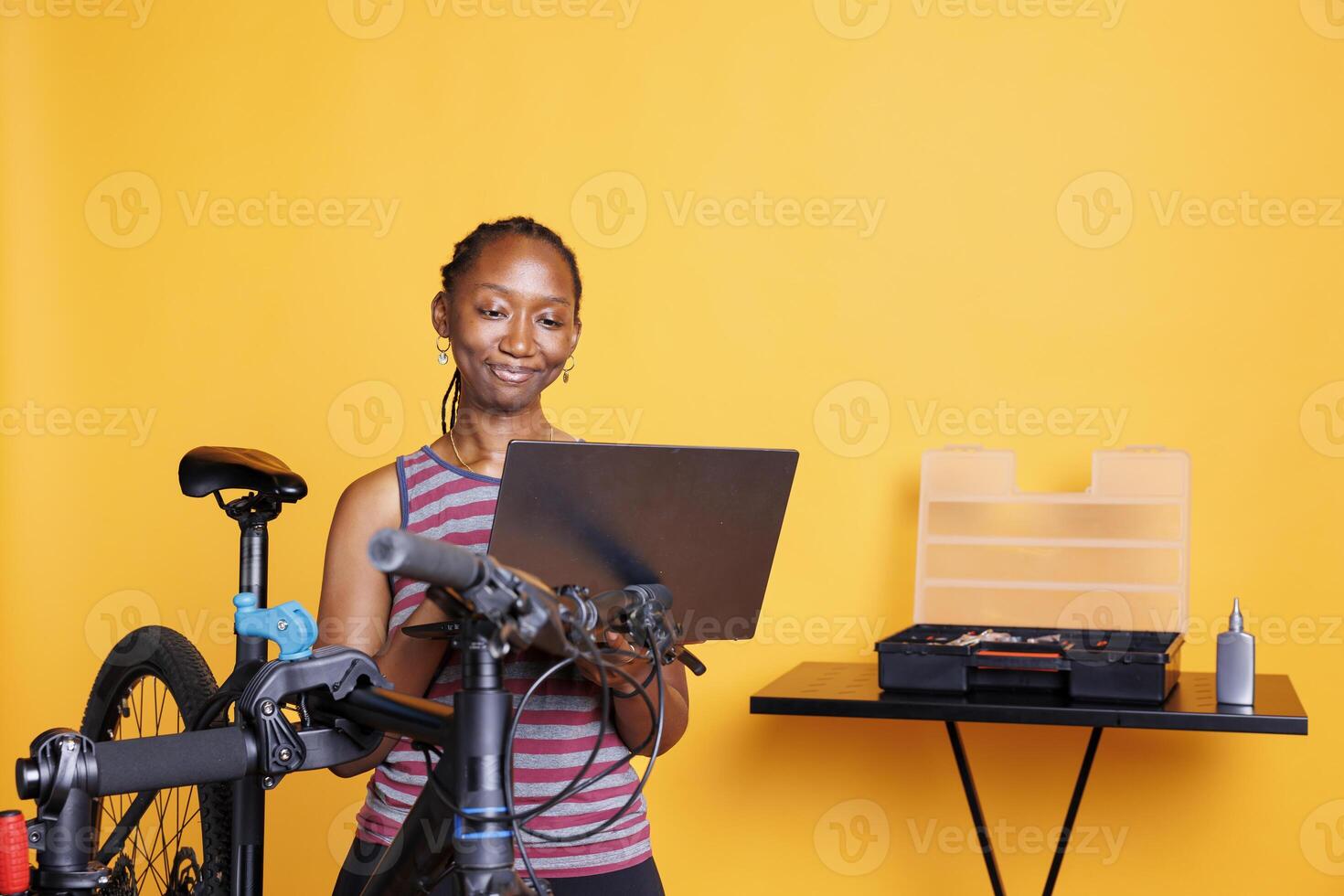 In front of yellow background, black woman performs bike maintenance by utilizing digital laptop for instructions. African american female using minicomputer and professional tools for fixing bicycle. photo
