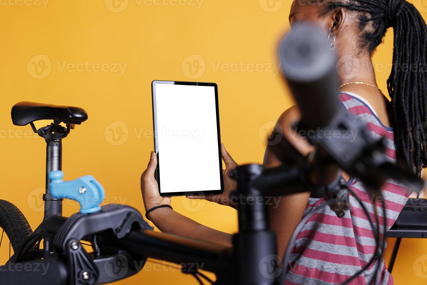 View in detail of african american female holding gadget with blank copyspace chromakey template and a nearby broken bicycle. Black woman using digital tablet with mockup whitescreen to maintain bike. photo