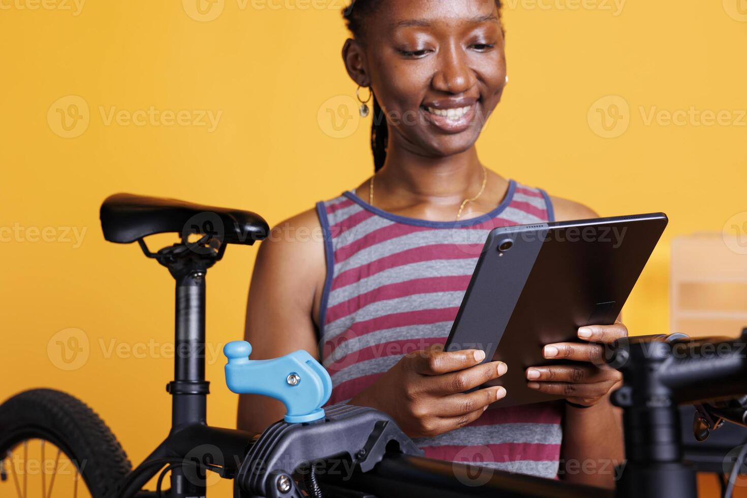 Energetic black woman carefully inspects and repairs bike frame using repair-stand and digital device. Image showing close-up view of female cyclist holding smart tablet while servicing bicycle. photo