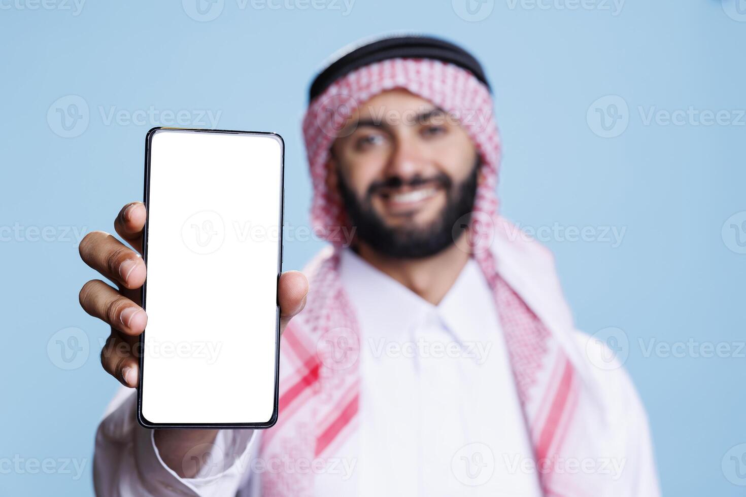 Man in arabic attire holding smartphone displaying white screen for application advertising. Smiling muslim person posing in studio and showing mobile phone blank touchscreen closeup photo
