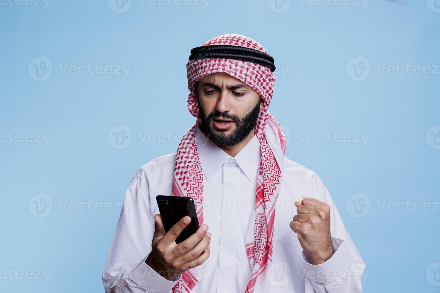 Excited muslim man wearing islamic traditional clothes reading news on social media using smartphone. Arab dressed in ghutra headscarf and thobe while checking message on mobile phone photo